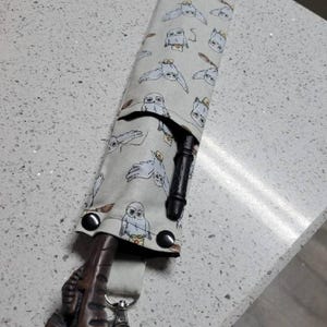 K&ouml;nnte beinhalten: Ein wei&szlig;er Stoff-Zauberstabhalter mit einem Muster aus Eulen. Der Halter hat einen schwarzen Gurt mit einem silbernen Clip. Ein brauner Holzstab ragt aus dem Halter.