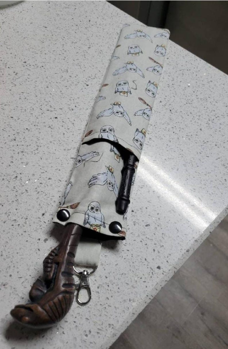K&ouml;nnte beinhalten: Ein wei&szlig;er Stoff-Zauberstabhalter mit einem Muster aus Eulen. Der Halter hat einen schwarzen Gurt mit einem silbernen Clip. Ein brauner Holzstab ragt aus dem Halter.