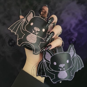 May include: Two black bat-shaped keychains with embroidered details. One bat has pink accents, the other has purple accents. Both have a cartoon-like design with wings and a smiling face. The keychains are held up against a dark purple background.