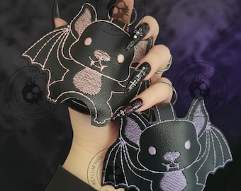 May include: Two black bat-shaped keychains with embroidered details. One bat has pink accents, the other has purple accents. Both have a cartoon-like design with wings and a smiling face. The keychains are held up against a dark purple background.
