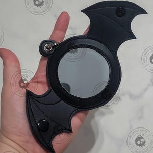 May include: A black bat-shaped compact mirror with a round mirror inside. The mirror has a black stitched border. The bat wings have a stitched design. The mirror has a metal ring for attaching a keychain.