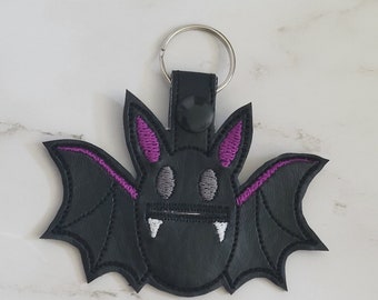 May include: A black bat-shaped keychain with purple ears and wings. The bat has white fangs and gray eyes. The keychain has a black leather strap and a silver metal ring.