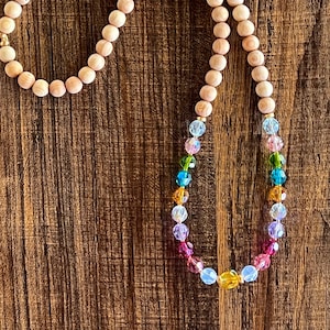 Puede incluir: Un collar largo de cuentas de madera con un arcoíris de cuentas de cristal. Las cuentas de madera son de color marrón claro y las cuentas de cristal son transparentes, rosas, amarillas, verdes, azules y moradas.