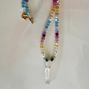 May include: A rainbow crystal necklace with a clear quartz point pendant and a gold heart charm. The necklace is made with small, faceted crystal beads in shades of red, orange, yellow, green, blue, and purple.