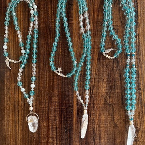 Op de afbeelding: Drie kristallen kettingen met heldere kwarts hangers. Elke ketting heeft een andere hanger: een vleugel, een ster en een halve maan. De kettingen zijn gemaakt van blauwe en heldere glazen kralen.