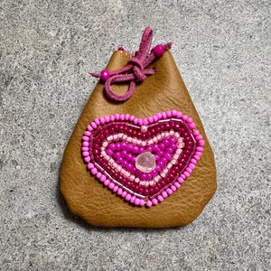 May include: A brown leather pouch with a beaded heart design in shades of pink and magenta. The heart features a central clear stone and is surrounded by a beaded border. The pouch has a drawstring closure with pink beads.