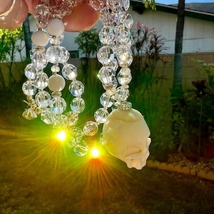 May include: A white skull pendant hangs from a crystal beaded necklace. The necklace is made of faceted crystal beads and white beads. The skull is carved from bone and has a detailed design. The necklace is photographed against a green grassy background.