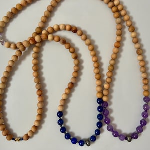 Op de afbeelding: Drie kettingen met houten kralen en edelsteenaccenten. Eén ketting heeft blauwe lapis lazuli kralen, een andere heeft paarse amethist kralen en de derde heeft een kleine cluster van paarse edelstenen. De houten kralen zijn lichtbruin.