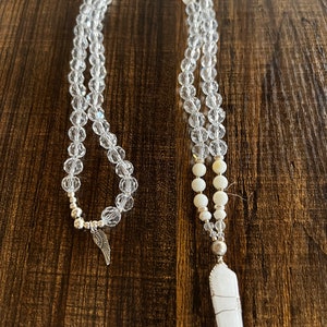 May include: A white crystal necklace with a clear crystal pendant and a silver angel wing charm. The necklace is made of clear crystal beads and white shell beads.