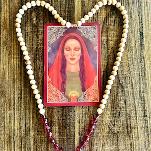 Puede incluir: Un collar de cuentas de madera en forma de corazón con una tarjeta roja que presenta a una mujer con los ojos cerrados en el centro. La tarjeta tiene un borde dorado y diseños intrincados. El collar tiene cuentas de cristal rojo en la parte inferior.