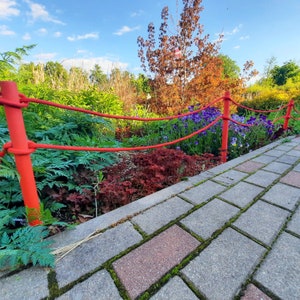 May include: A red rope fence with wooden posts, separating a brick pathway from a garden with green foliage and purple flowers.
