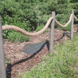 May include: A rope fence made of natural rope strung between wooden posts. The fence is in a garden setting with green grass and brown mulch.