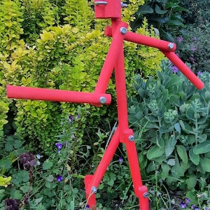 May include: A red wooden stick figure sculpture with a human-like face and arms outstretched. The sculpture is standing in a garden with green plants and purple flowers.