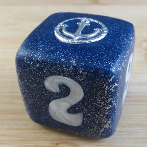 May include: A navy blue dice with white numbers and an anchor symbol. The dice has a textured surface and is resting on a wooden surface. The number "2" is visible on one side.