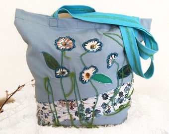 May include: A light blue tote bag with a floral design. The bag features embroidered white and brown flowers with green leaves and stems. The handles are a bright turquoise color.