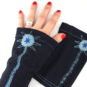 Könnte beinhalten: Ein Paar dunkelmarineblaue fingerlose Handschuhe aus strukturiertem Stoff. Jeder Handschuh zeigt ein gesticktes hellblaues, kreisförmiges Blumenmotiv mit strahlenden Blütenblättern und einem langen, dekorativen Stiel in türkisfarbenem Garn.