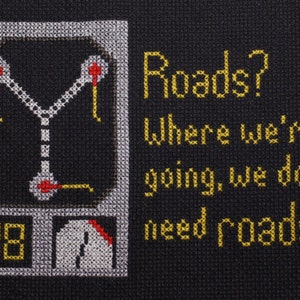 May include: Cross-stitch art on black fabric featuring a DeLorean time machine schematic and the quote "Roads? Where we're going, we don't need roads." The design includes yellow, white, red, and black thread, creating a retro, geeky aesthetic.