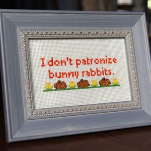 Puede incluir: Un bordado de punto de cruz enmarcado en gris con el texto "No patrocino conejos de peluche." El bordado presenta cinco conejos marrones con orejas naranjas y flores amarillas.