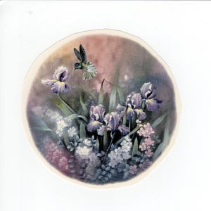 Puede incluir: Un plato decorativo redondo con una pintura de acuarela de lirios y otras flores en tonos morados, blancos y rosas. Un colibrí se cierne sobre las flores. El plato tiene un borde color crema.