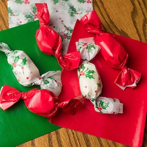 May include: Four small white candies wrapped in red and white patterned paper with a holly design. The candies are on a green and red background.