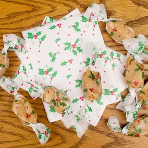 May include: A collection of individually wrapped peanut butter candies, each wrapped in white paper with a green holly leaf and red berry pattern. The candies are arranged on a wooden surface.