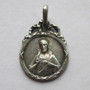 May include: A silver-toned pendant featuring a detailed depiction of a religious figure within an oval frame. The pendant has a loop at the top for a chain and is surrounded by decorative elements, including a ribbon and a laurel wreath.