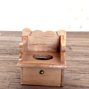 May include: A miniature wooden chair with a scalloped backrest and a circular cutout in the seat. The piece features a light brown wood grain finish and a small, centered brass-colored knob on the front panel below the seat, showing a compact design.