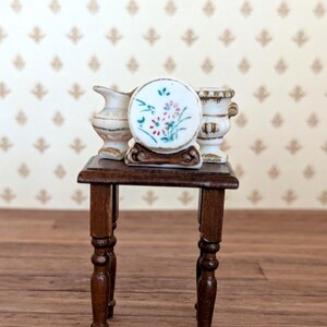 May include: A miniature dark brown wooden table with four turned legs, supporting a small white ceramic pitcher, a round white ceramic plate with a red and green floral pattern on a brown stand, and a white ceramic urn with gold-toned trim.