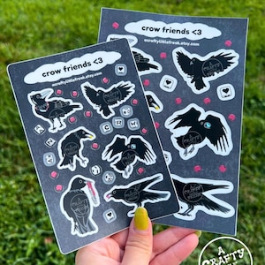 Puede incluir: Dos hojas de pegatinas con ilustraciones de cuervos en varias poses, con el texto "crow friends <3" y "acraftylittlefreak.etsy.com". Las pegatinas son en blanco y negro con detalles en rosa y pequeños gráficos de corazón y dados.