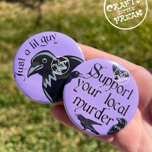 Puede incluir: Dos botones redondos morados con ilustraciones de cuervos y texto blanco. Uno dice "Just a lil guy" y el otro "Support your local murder". Ambos tienen un logo de "Crafty Little Freak".