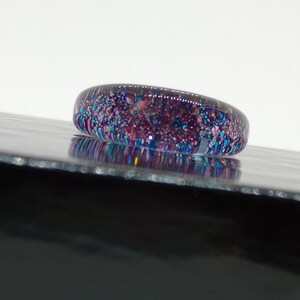 Puede incluir: Un anillo con una banda de vidrio transparente llena de purpurina iridiscente en tonos rosa, azul y morado.