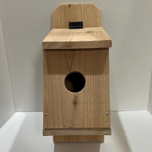 Pode incluir: Uma casa de pássaros de madeira com um telhado articulado e um buraco de entrada redondo. A casa de pássaros é feita de madeira natural e tem um aspecto rústico.