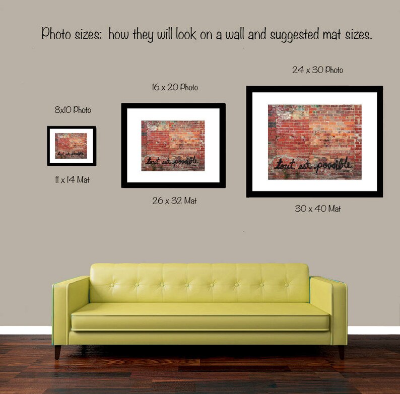 May include: Three framed photos of different sizes showing a brick wall with the text "tout est possible" written in black paint. The photos are hanging on a light brown wall above a yellow couch.