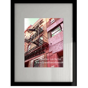 May include: Framed photograph of a brick building with a fire escape. The building is a mix of red and tan bricks, with red trim around the windows and fire escape. The photograph is in a black frame with a white mat.