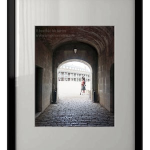 Puede incluir: Un arco de ladrillo conduce a un patio con una persona con un uniforme rojo y blanco de pie en el centro. El texto "© heather luk berios www.greenhoneydew.com" es visible en la esquina superior izquierda de la imagen.