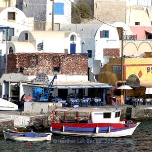 May include: A white boat with red and blue trim is docked at a waterfront in a Greek village. The boat has a small cabin and a wooden deck. The village is built on a hillside and has whitewashed buildings with blue accents. There are several restaurants and shops in the village, including one with a sign that says "Sunset Taverna".
