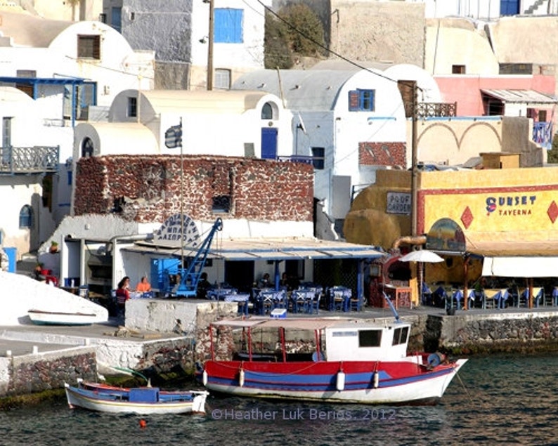 May include: A white boat with red and blue trim is docked at a waterfront in a Greek village. The boat has a small cabin and a wooden deck. The village is built on a hillside and has whitewashed buildings with blue accents. There are several restaurants and shops in the village, including one with a sign that says "Sunset Taverna".