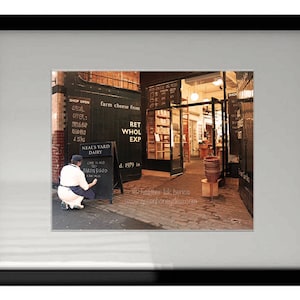 Peut inclure: Une personne portant une chemise blanche et un pantalon bleu se tient devant un tableau noir sur lequel est inscrit "Neal's Yard Dairy" et "Farm cheese from". Le panneau est dans un magasin avec un sol en bois et une porte menant à une autre pièce.