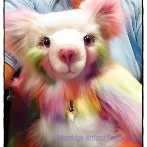 May include: A white teddy bear with rainbow fur and blue eyes. The bear is wearing a gold necklace with a heart pendant. The bear is being held by a person wearing a light blue shirt. The text "Blue Ridge Artisan Teddy Bears" and "Mindy Sharlow" is visible in the image.