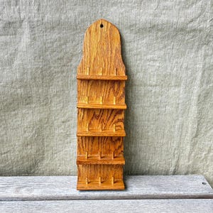 May include: A wooden spice rack with four shelves and a decorative top. The rack is made of light brown wood and has a hole at the top for hanging. The shelves are designed to hold small jars or containers. The rack is displayed on a wooden surface.