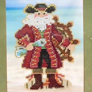 May include: Cross-stitch design of a pirate with a white beard, wearing a red coat, black pants, and a tricorn hat. The pirate is holding a map and a ship's wheel. The background shows a beach and blue sky.