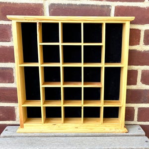 May include: A wooden display shelf with multiple square compartments. The shelf is made of light-colored wood and has a black backing. It is designed to hold small items and is set against a brick wall.