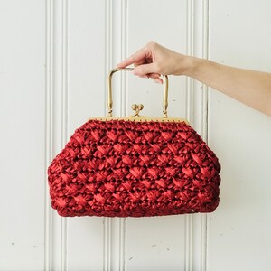 May include: A vintage red handbag with a woven texture and a gold-colored metal frame. The bag has a clasp closure and a handle. The bag is held up by a hand.