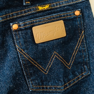 Peut inclure: Gros plan sur la poche arri&egrave;re d'un jean bleu fonc&eacute;. La poche pr&eacute;sente un motif en 'W' cousu et un patch en cuir marron avec le logo 'Wrangler'. Les jeans ont des rivets en cuivre et des coutures jaunes.