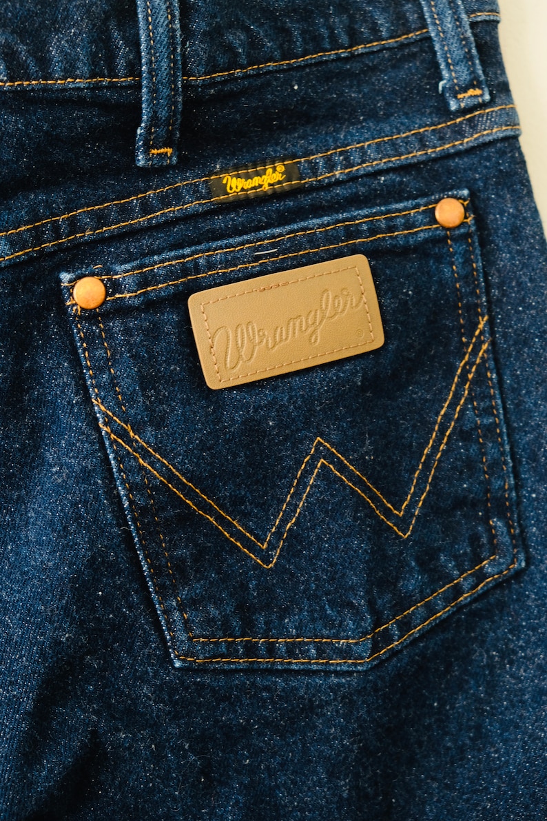 Peut inclure: Gros plan sur la poche arri&egrave;re d'un jean bleu fonc&eacute;. La poche pr&eacute;sente un motif en 'W' cousu et un patch en cuir marron avec le logo 'Wrangler'. Les jeans ont des rivets en cuivre et des coutures jaunes.
