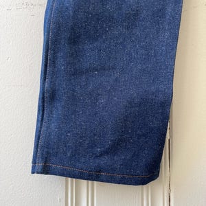 May include: Close-up of a section of blue denim jeans. The fabric has a textured appearance with visible threads. The hem is finished with a contrasting orange-brown stitch. The jeans are hanging against a white background.
