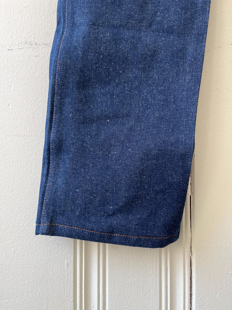 May include: Close-up of a section of blue denim jeans. The fabric has a textured appearance with visible threads. The hem is finished with a contrasting orange-brown stitch. The jeans are hanging against a white background.