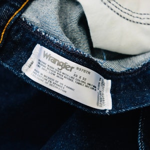 Peut inclure: Gros plan sur un jean Wrangler bleu fonc&eacute;, montrant les d&eacute;tails int&eacute;rieurs. L'&eacute;tiquette indique "Wrangler 937STR 33 x 32" avec les instructions d'entretien et la composition du tissu. Le jean est fabriqu&eacute; au Mexique.