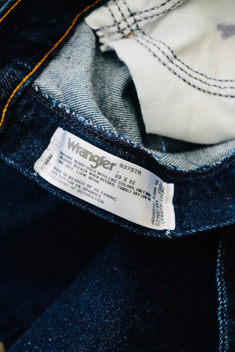 Peut inclure: Gros plan sur un jean Wrangler bleu fonc&eacute;, montrant les d&eacute;tails int&eacute;rieurs. L'&eacute;tiquette indique "Wrangler 937STR 33 x 32" avec les instructions d'entretien et la composition du tissu. Le jean est fabriqu&eacute; au Mexique.