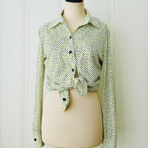 May include: A long-sleeved button-down shirt on a mannequin. The shirt is white with a pattern of small green and navy blue dots. The shirt is tied at the waist and has navy blue buttons and cuffs. The mannequin is on a metal stand.